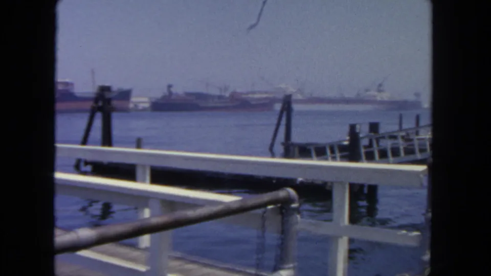 1962 harbor footage from a dock in San Diego, showing foreground railings, pilings, blue water, and tankers beyond.