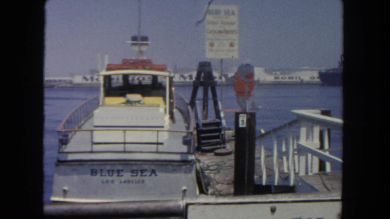1962 8mm frame of the Blue Sea boat at a Southern California dock, with a ferry sign, life ring, pilings, and harbor traffic in the background.