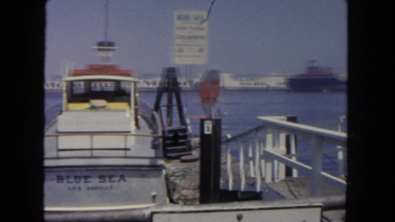 The Blue Sea remains docked in this 1962 harbor view, framed by pier railings, a ferry sign, a life ring, and distant ships.