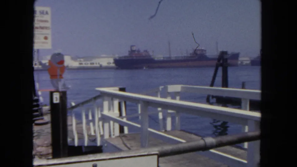 The camera pans away from the Blue Sea toward white dock railings, a partial ferry sign, calm harbor water, and a cargo vessel in the distance.