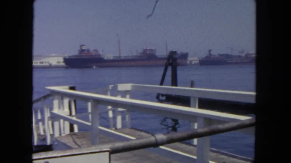 Final 1962 harbor frame showing white pier railings and calm water, with large vessels across the harbor and no people visible.