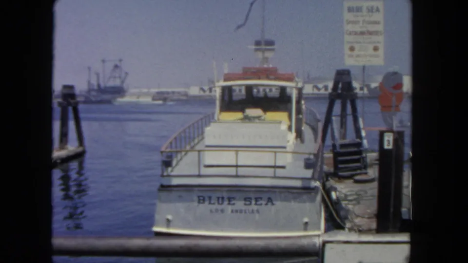 1962 San Diego harbor frame showing the Blue Sea docked near a sign, with industrial waterfront details across the water.