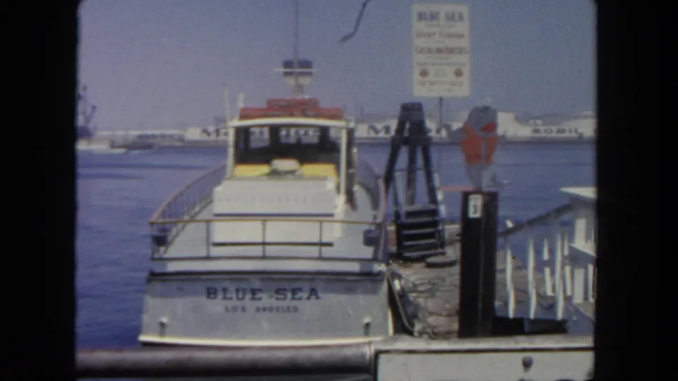 Final 1962 San Diego dockside frame of the Blue Sea boat, a pier sign, gangway, calm water, and distant harbor structures.