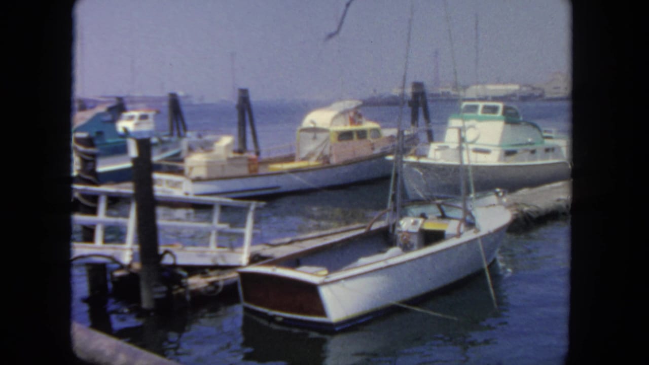 1962 San Diego marina view with docked small boats, wooden pilings, hazy water, and distant harbor structures.