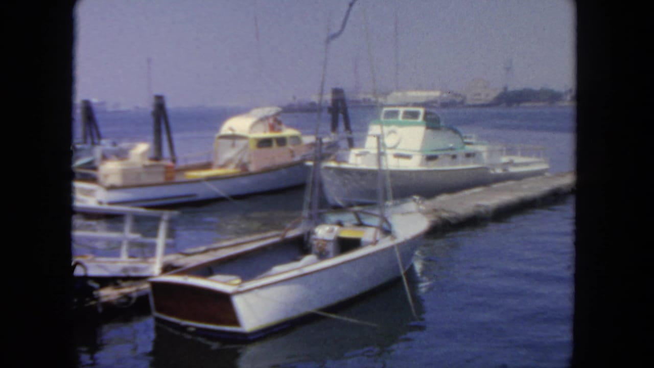 1962 San Diego harbor frame showing a small sailboat in front of docked motorboats and wooden pier structures.