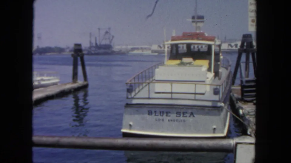 1962 San Diego harbor frame with the BLUE SEA LOS ANGELES boat stern, marina pilings, calm water, and distant waterfront industry.