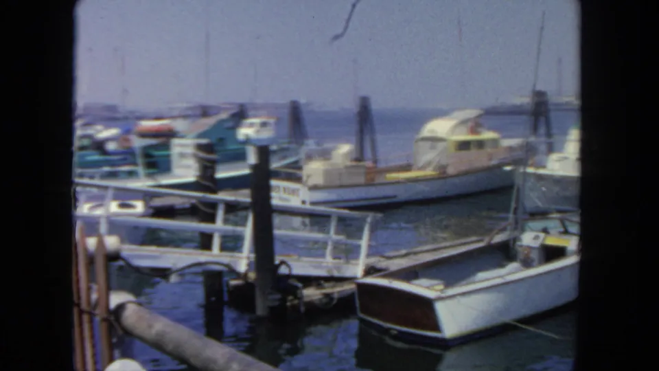 1962 San Diego marina view with a white foreground boat, railings, pilings, moored cabin cruisers, and calm harbor water.
