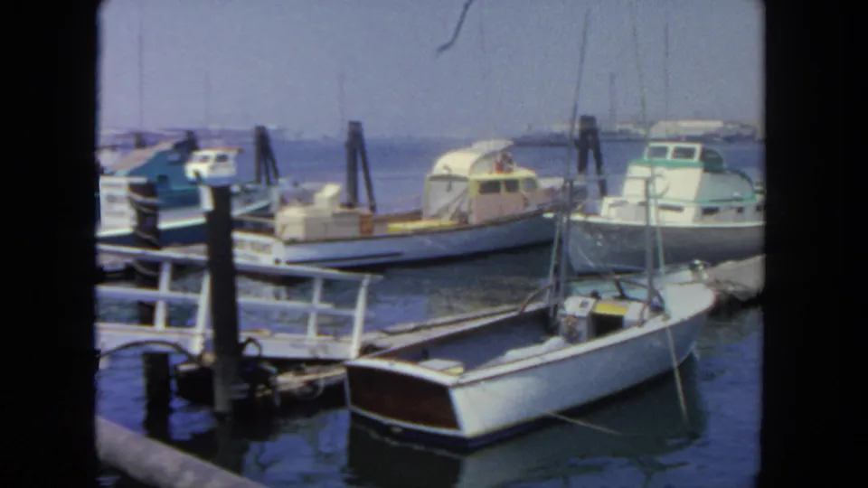 1962 San Diego harbor frame with multiple tied boats, a white foreground vessel, dock railings, pilings, calm water, and hazy background coast.