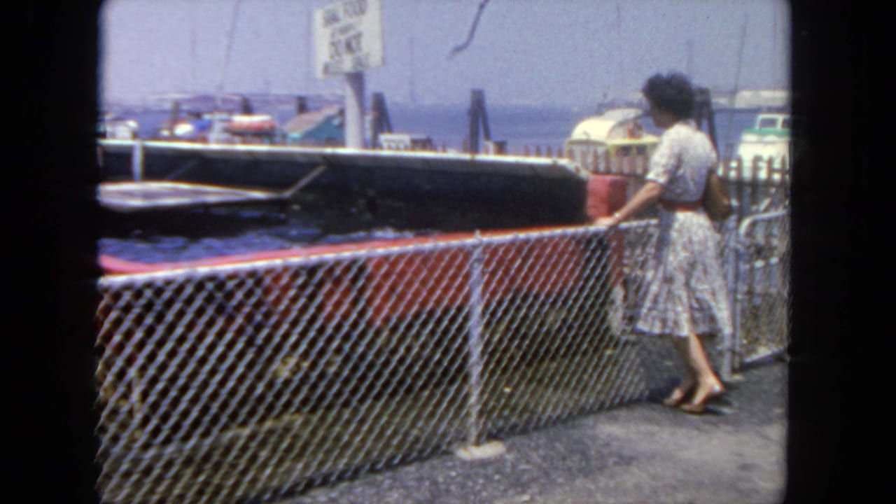 In 1962 San Diego home-movie footage, a woman stands beside a chain-link fence looking toward moored boats and water.