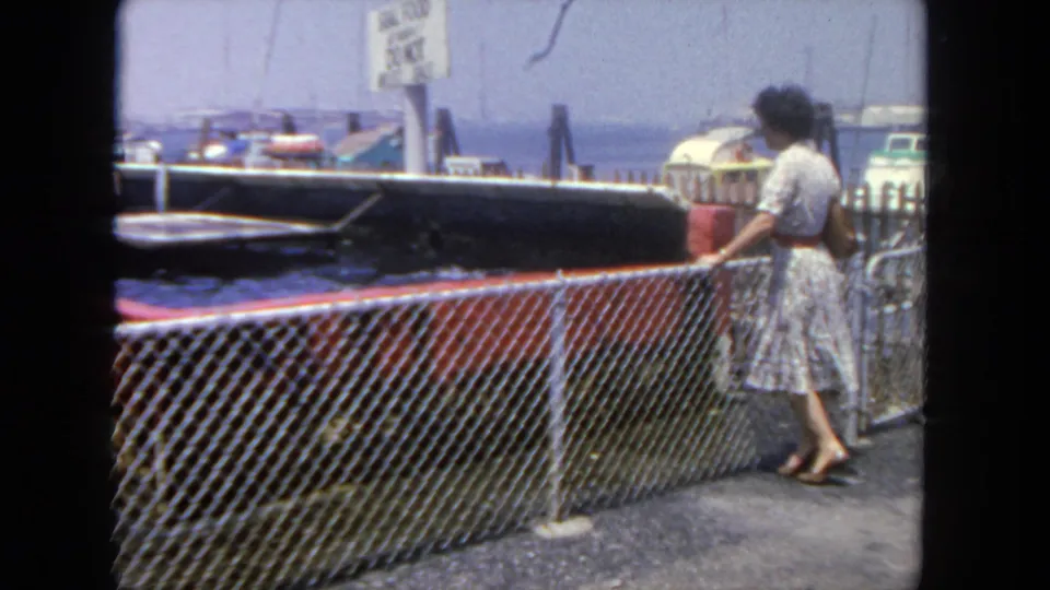In 1962 San Diego home-movie footage, a woman stands beside a chain-link fence looking toward moored boats and water.