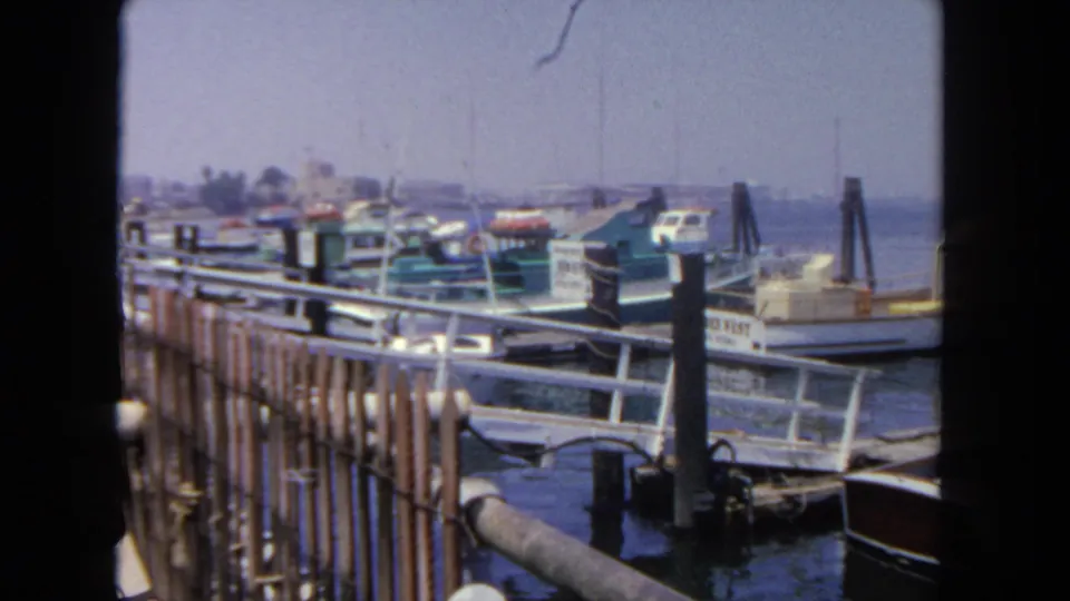 The view shifts to San Diego harbor boats, dock railings, vertical pilings, water, and a hazy distant shoreline in 8mm footage.