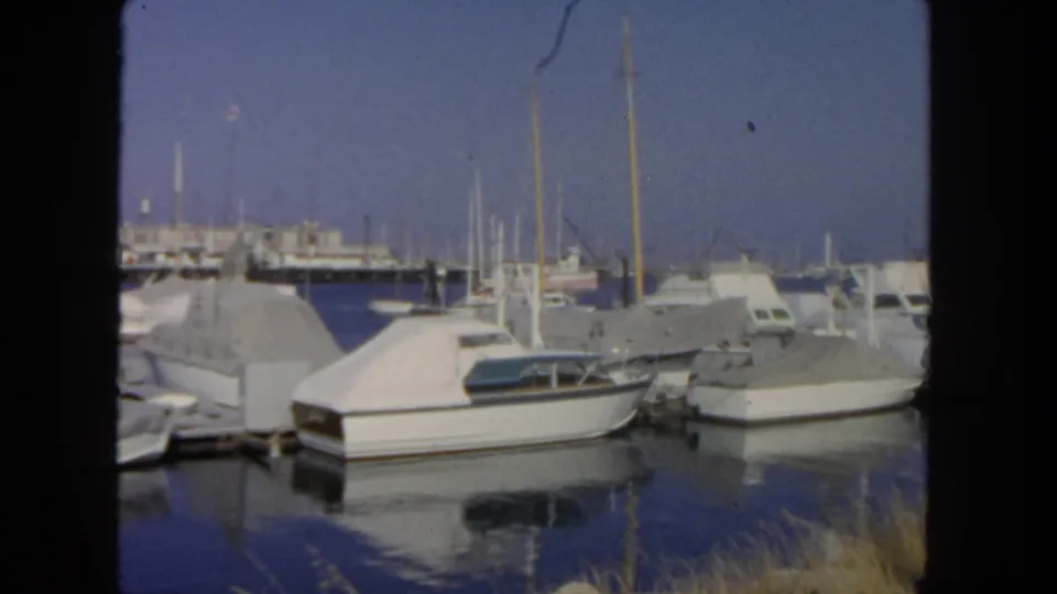 1962 home-movie frame of a San Diego harbor dock with covered cabin boats, small craft, masts, and shoreline details.