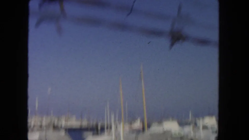 Blurred final 1962 San Diego harbor frame with mast tops, docked boats, blue sky, and dark overhead wires crossing the view.