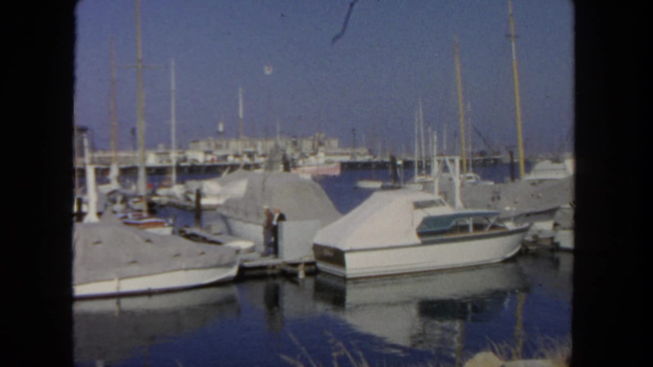 1962 San Diego marina view with covered motorboats, sailboat masts, calm water, and a man standing on the dock.