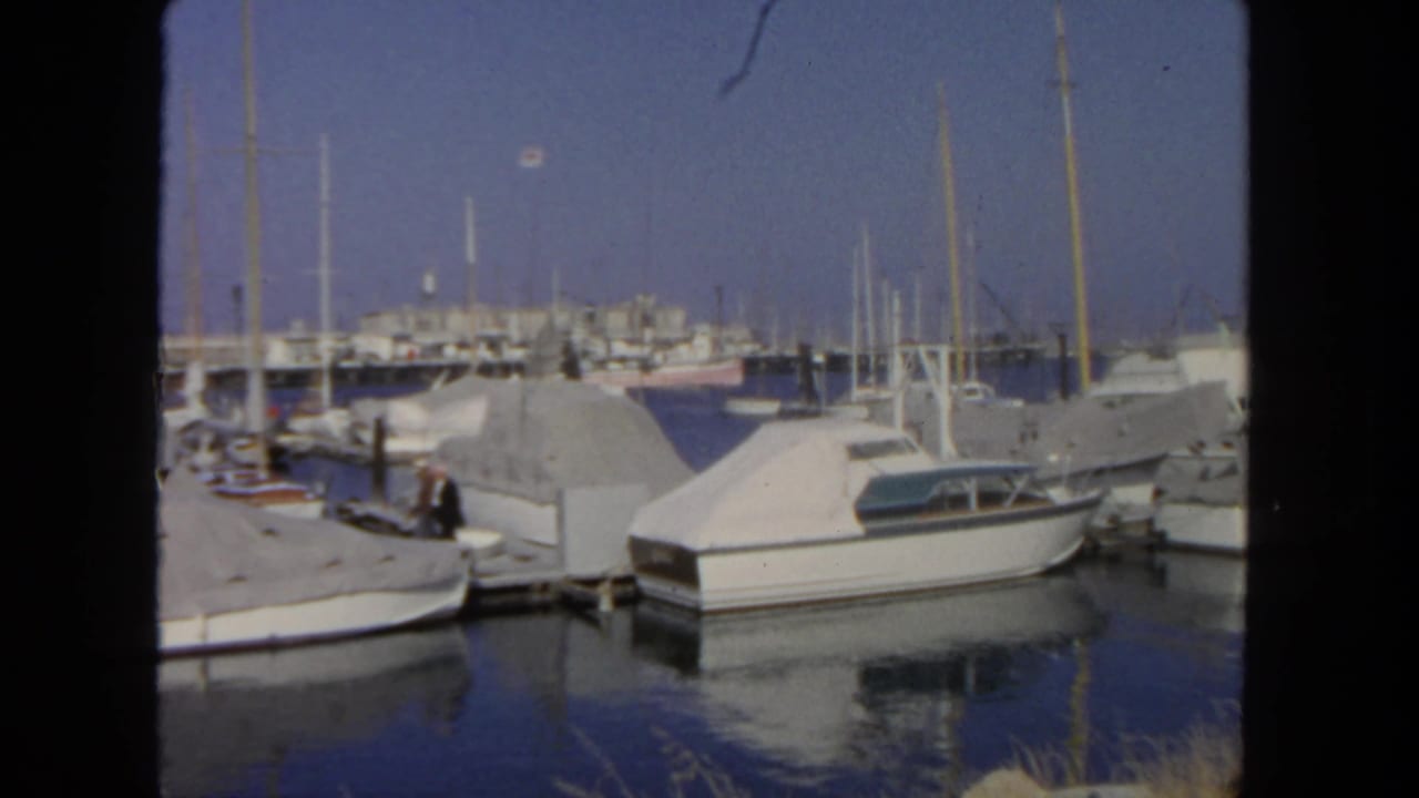 1962 San Diego marina scene showing white boats under covers, dock pilings, reflections, and a person near the dock.