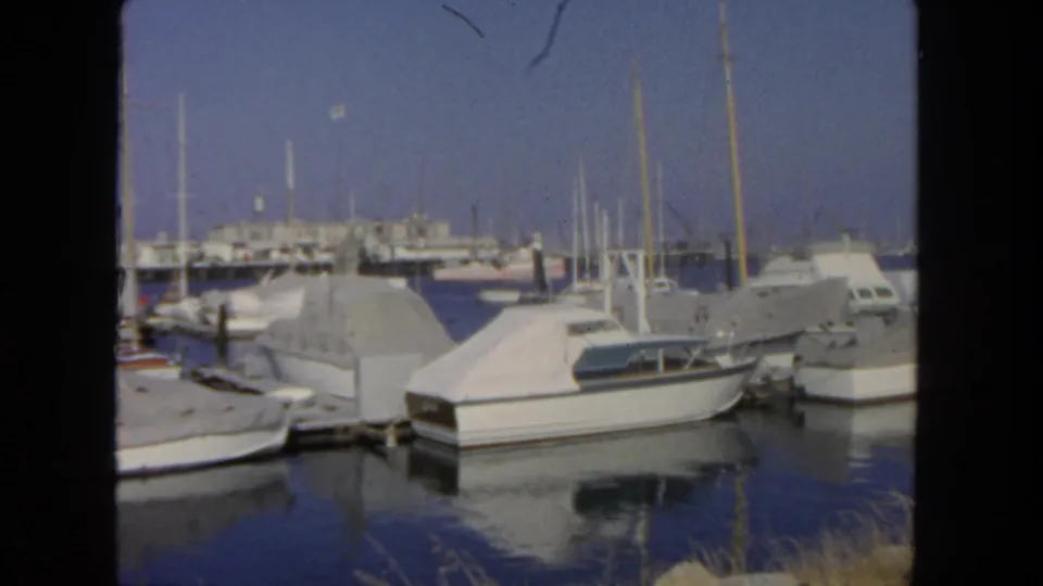 1962 San Diego marina view with covered pleasure boats, tall masts, dock pilings, reflections, and distant waterfront buildings.