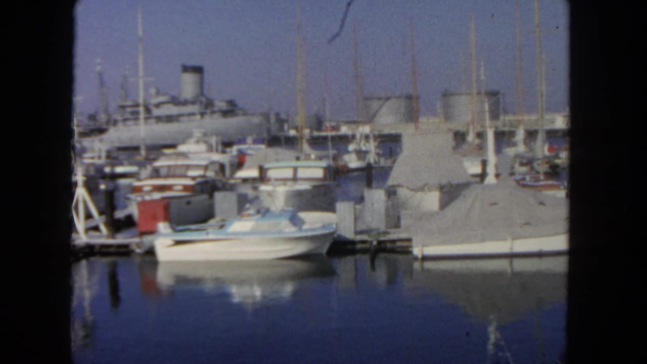 1962 San Diego marina view with moored pleasure boats, covered slips, mast lines, reflected water, and a distant large passenger ship.