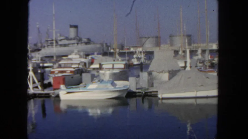 1962 San Diego marina view with moored pleasure boats, covered slips, mast lines, reflected water, and a distant large passenger ship.