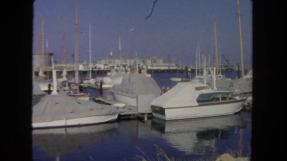 1962 San Diego marina scene with sailboats, covered moorings, a cabin cruiser, calm water, and indistinct people on distant docks.