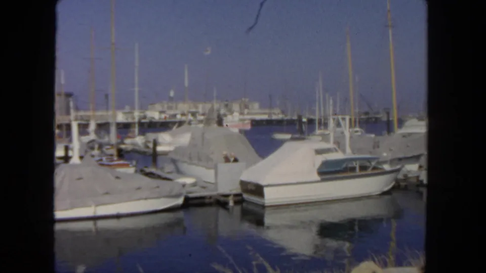 San Diego marina frame with a cabin cruiser in the foreground, sailboat masts, covered slips, water reflections, and indistinct dockside figures.