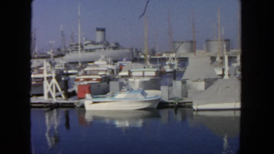 San Diego marina view from 1962 with a white motorboat, covered craft, dock pilings, masts, and a large liner in the background.