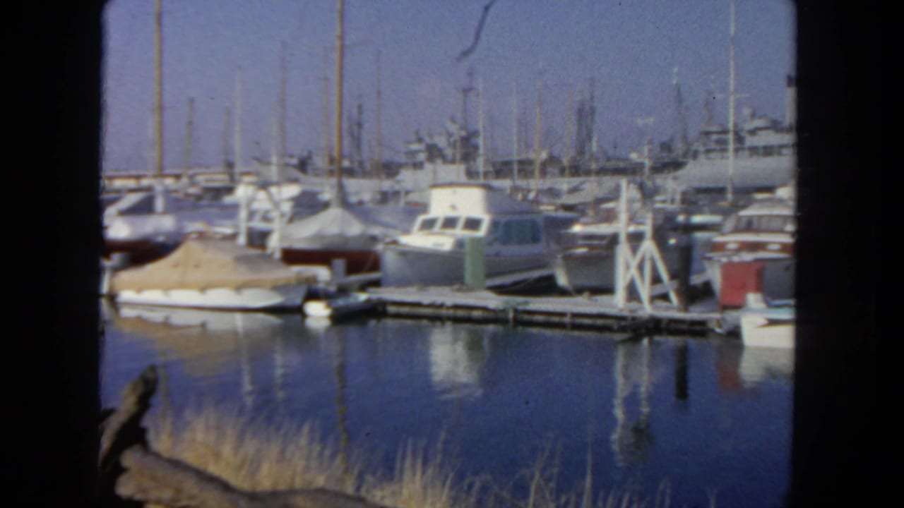 Small boats docked in a marina with blue water reflections and a larger vessel behind them.