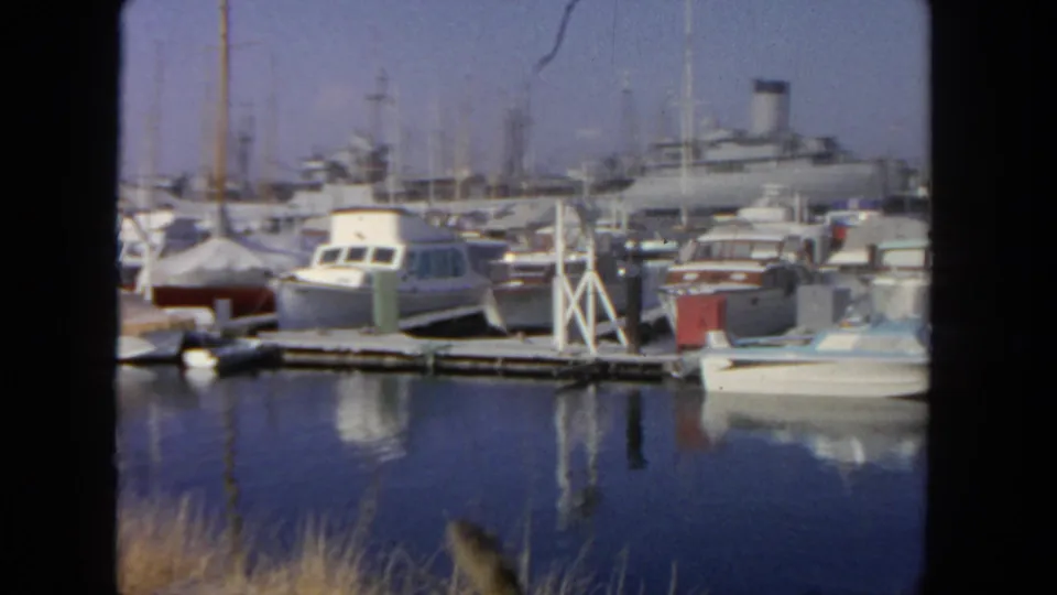1962 San Diego marina footage with white motorboats, sailboat masts, a wooden pier, shoreline grass, and a large vessel in back.