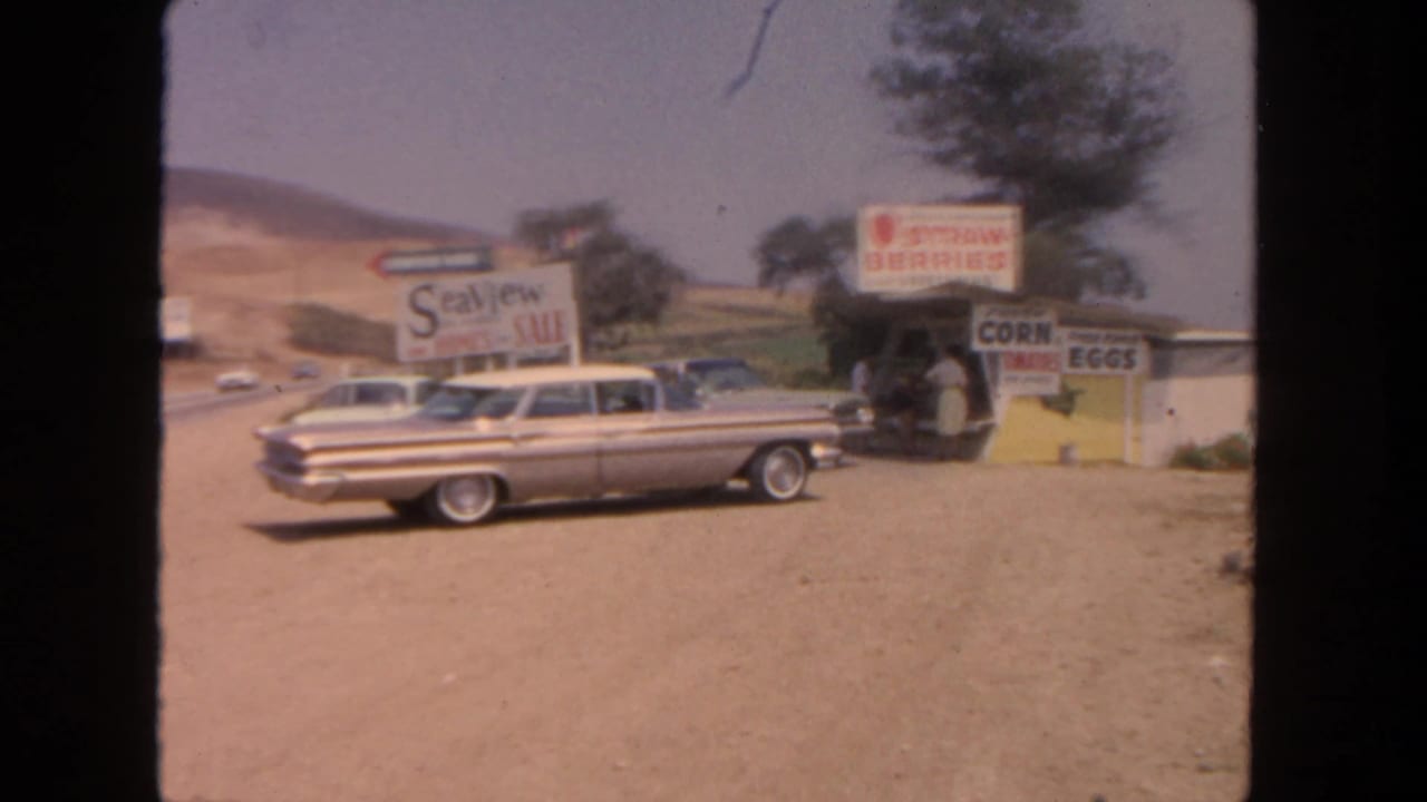 1962 San Diego-area roadside stand with strawberry, corn, and eggs signs beside a parked two-tone sedan.