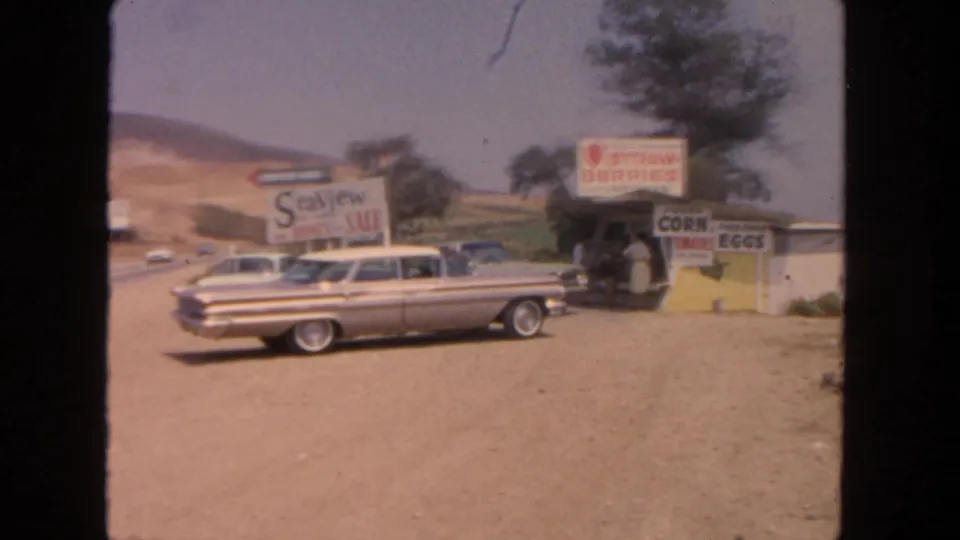 1962 San Diego-area roadside stand with strawberry, corn, and eggs signs beside a parked two-tone sedan.