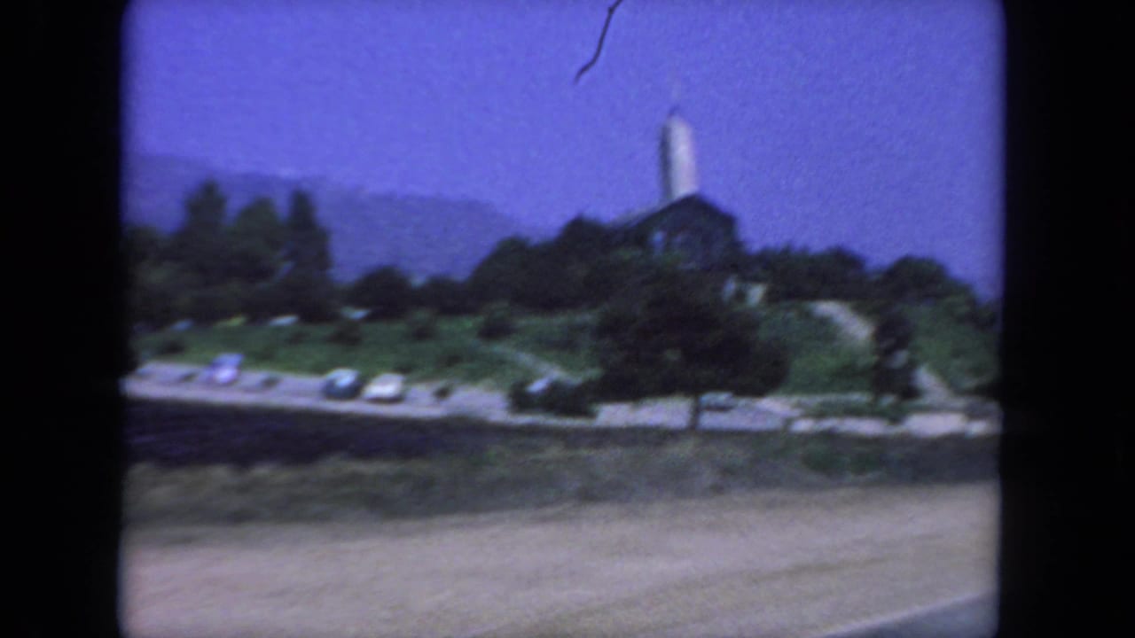 1962 San Diego home-movie frame showing a hilltop church with a tall steeple, trees, road edge, and distant hills.