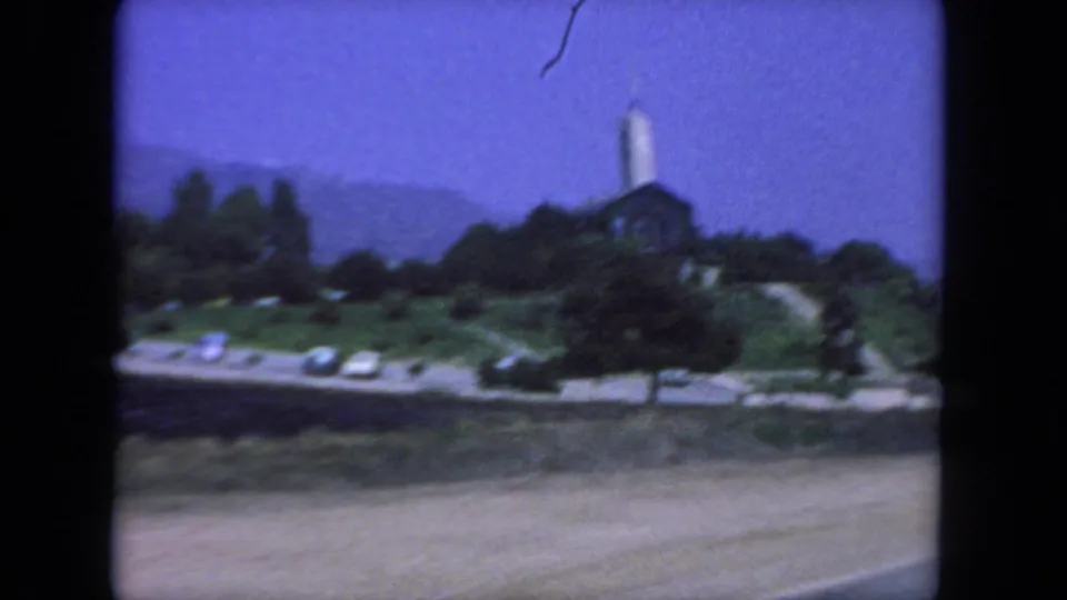 1962 San Diego home-movie frame showing a hilltop church with a tall steeple, trees, road edge, and distant hills.