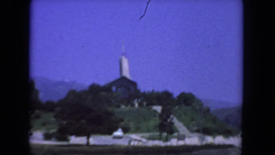 1962 San Diego driving-view frame with a church and steeple on a landscaped hill under a deep blue sky.