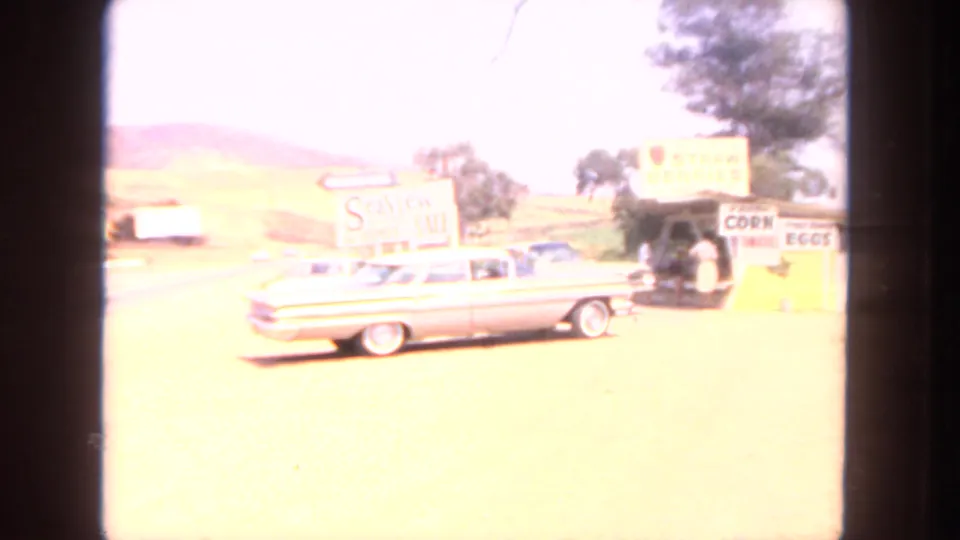 1962 San Diego roadside frame showing a white sedan near a produce stand with food signs and small figures nearby.