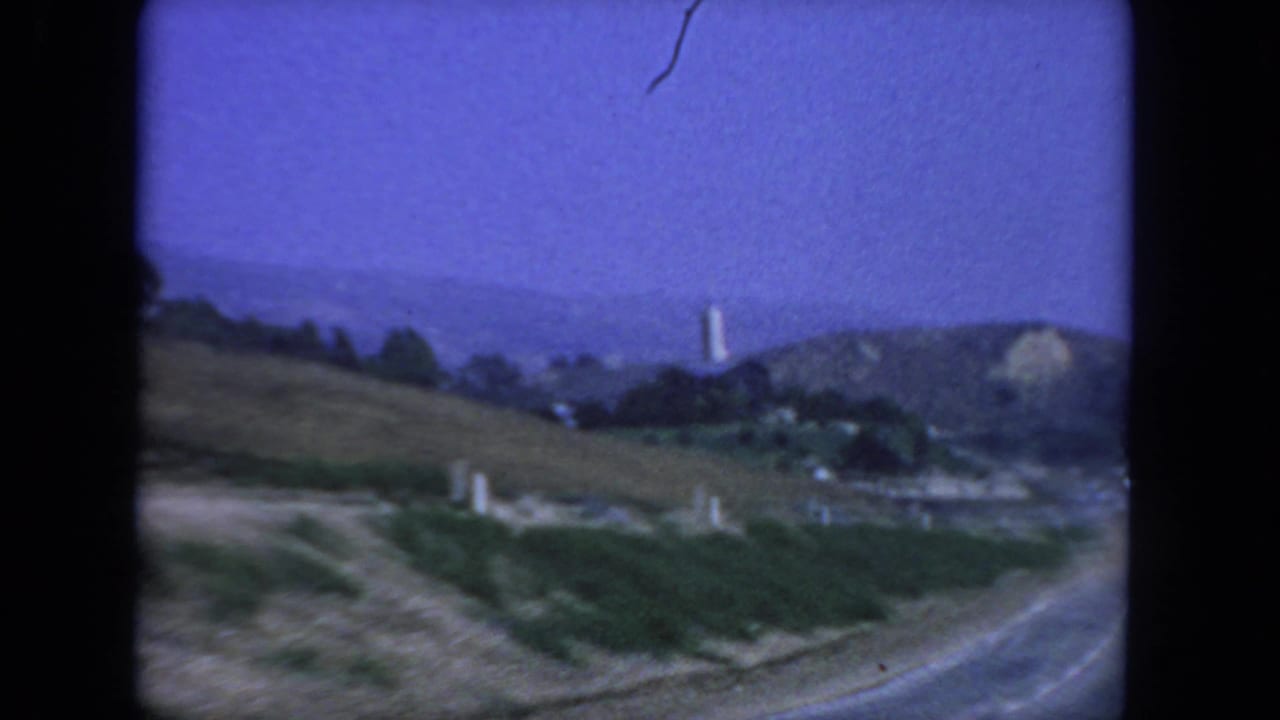 Road edge and hillside vegetation with distant ridges and a pale vertical tower form.
