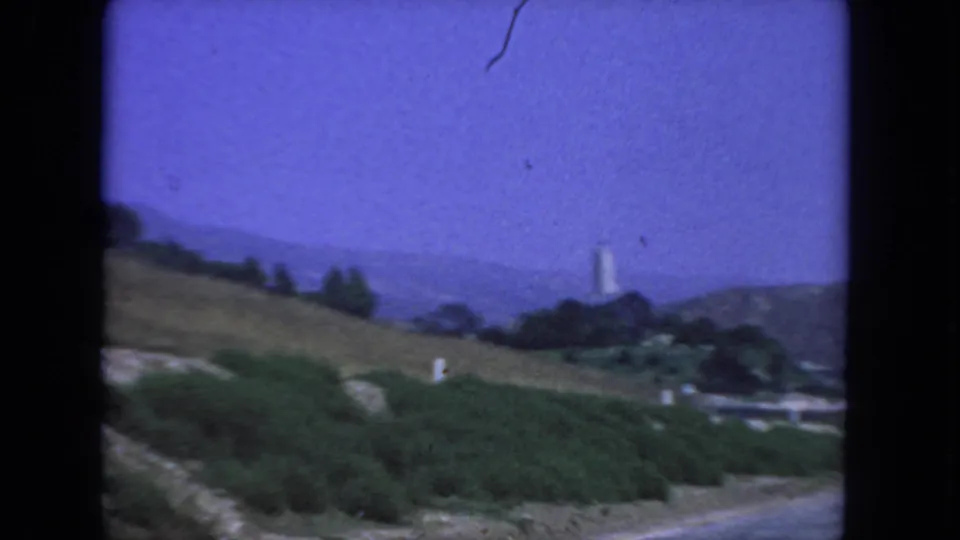 Final 1962 8mm frame of San Diego-area roadside hills, brush, distant trees, and a pale tower-like structure under a blue-purple sky.