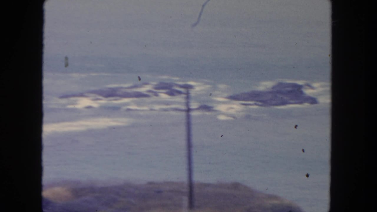 1962 San Diego coastal view from a roadside position, showing rocky surf, muted sky, and a utility pole crossing the frame.