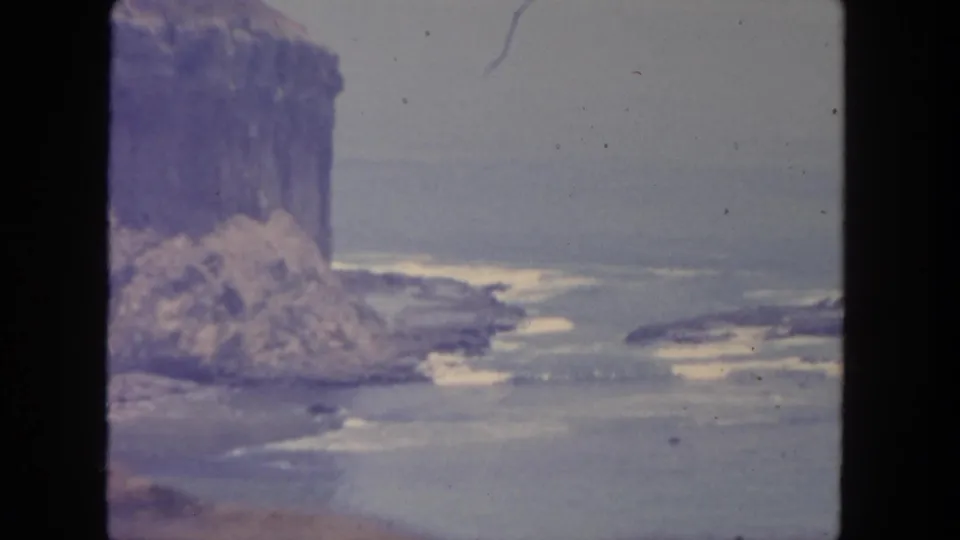 1962 San Diego shoreline view with a bluff at left, rocky surf below, and soft home-movie exposure.