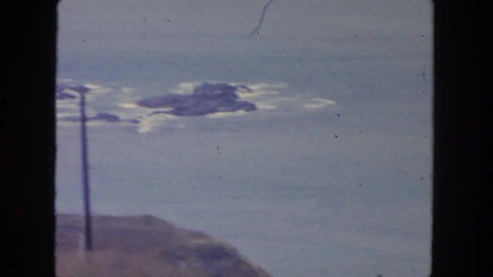 Vintage 8mm San Diego coastal view with rocky shoreline, small offshore rocks, calm water, and a foreground utility pole.