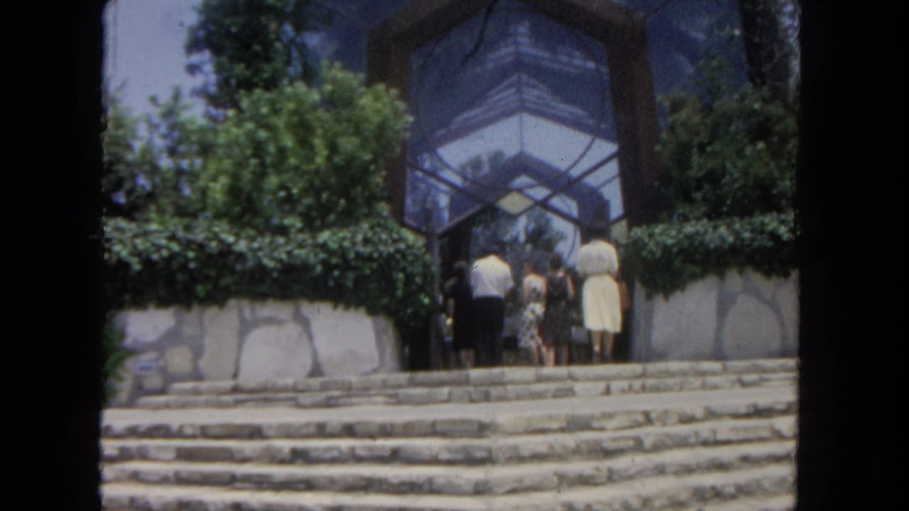 In 1962 San Diego home-movie footage, adults in period clothing approach a geometric glass atrium above stone steps.