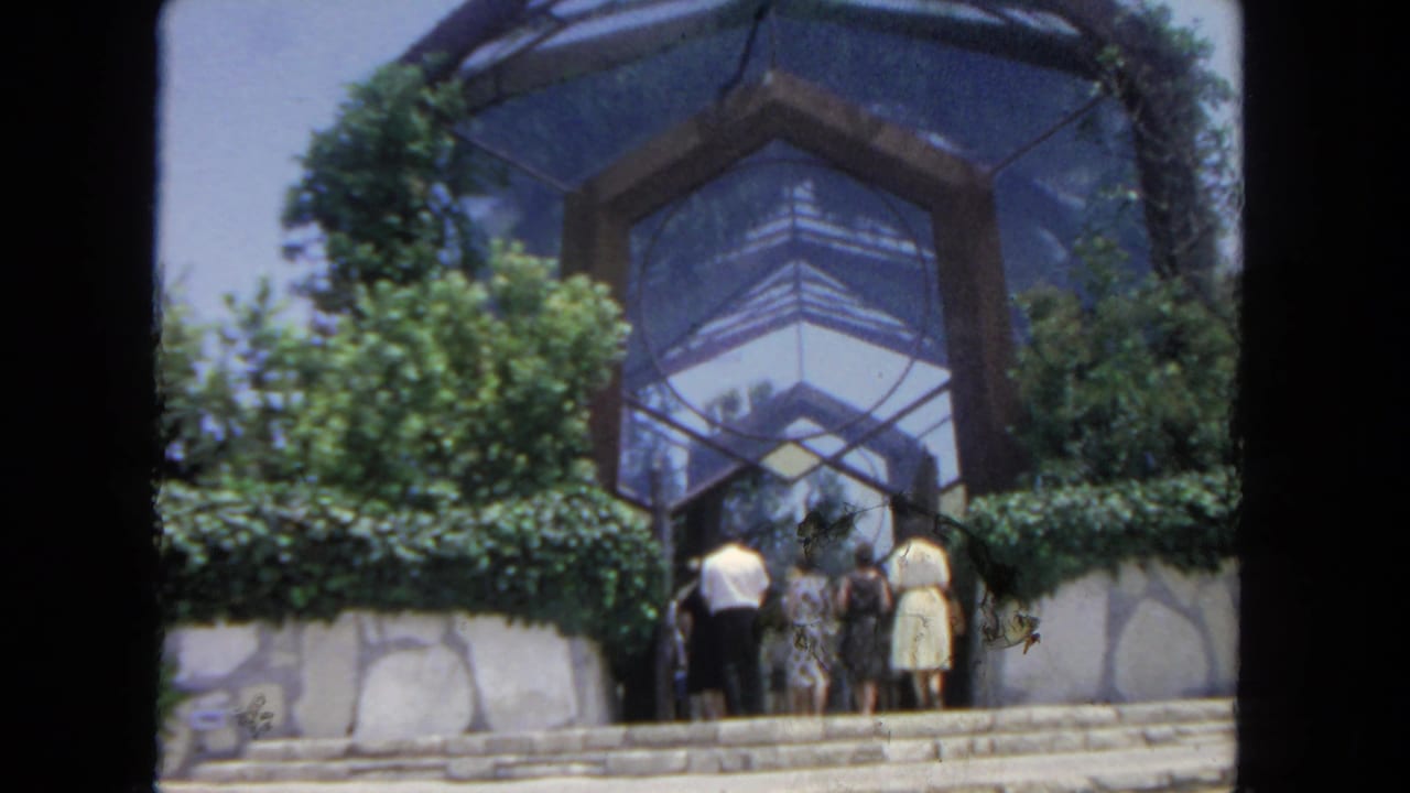A small group in 1960s attire gathers by the glass atrium entrance, with hedges and stonework around them.