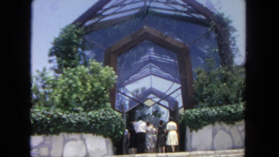 Visitors in 1960s clothing move toward the angular glass atrium, framed by shrubs and a low stone wall.