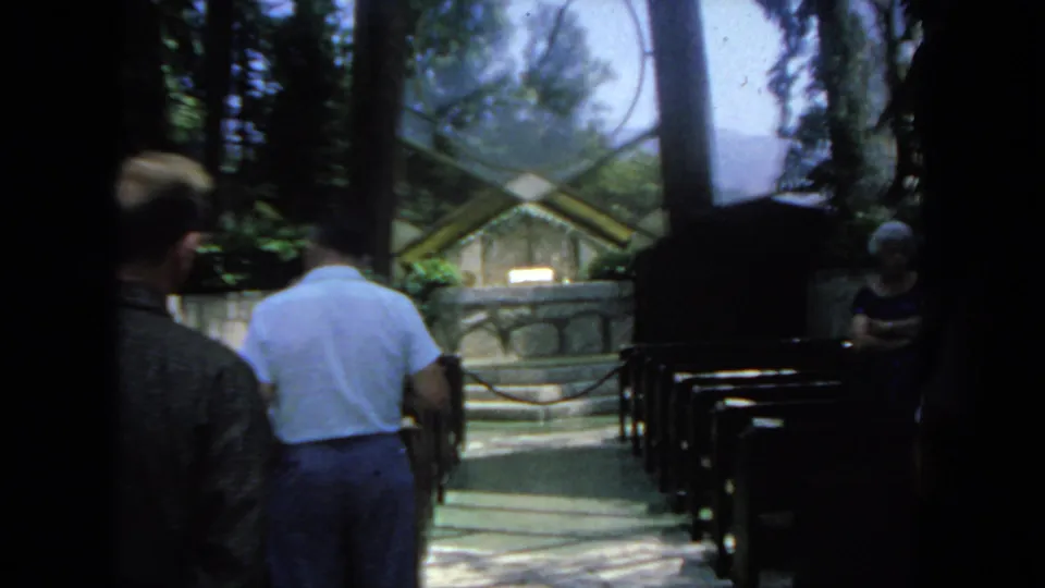Inside the San Diego chapel-like space, visitors walk past wooden pews toward a stone altar area beneath trees and glass.