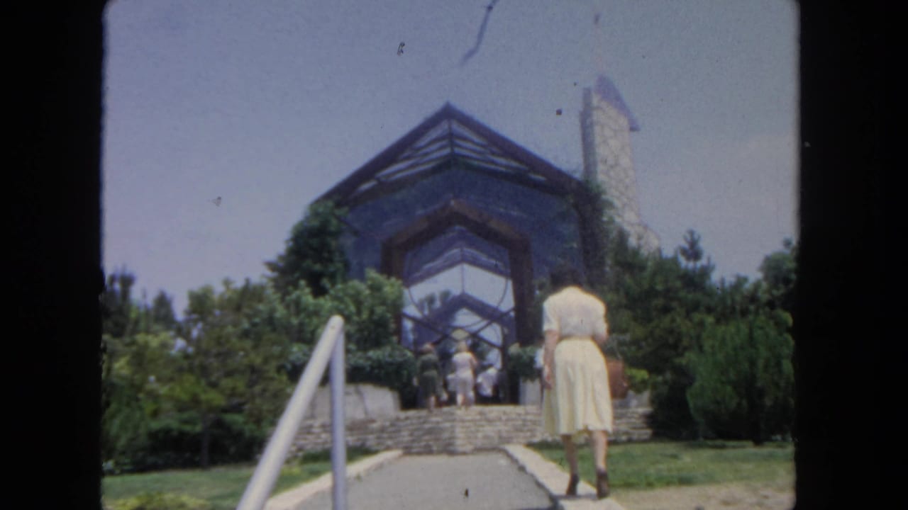 In 1962 San Diego, a woman in a pale yellow dress approaches stone steps leading to a landscaped glass-and-stone building.