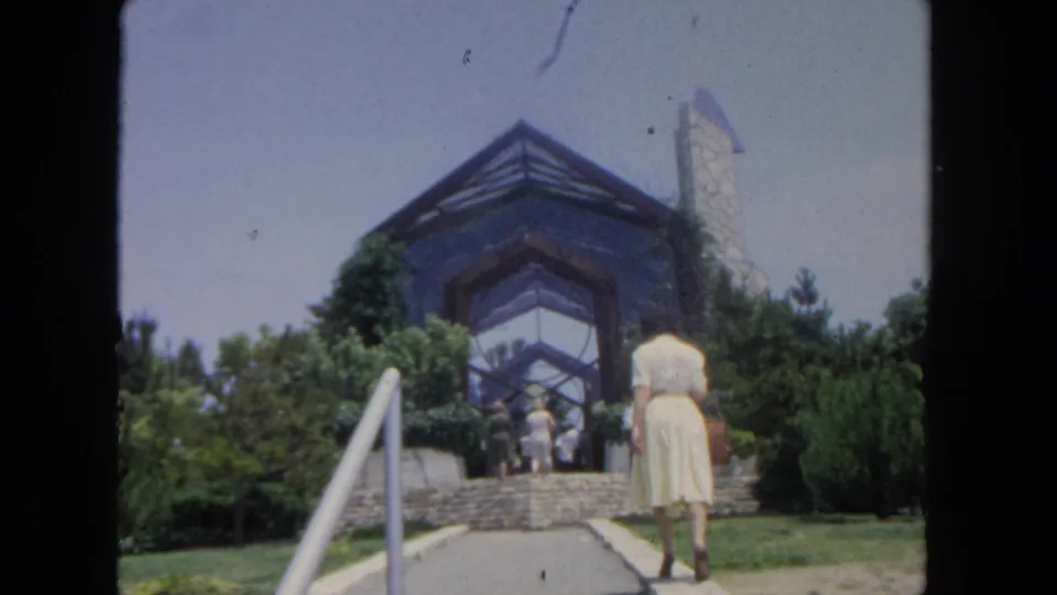 In 1962 San Diego, a woman in a pale yellow dress approaches stone steps leading to a landscaped glass-and-stone building.