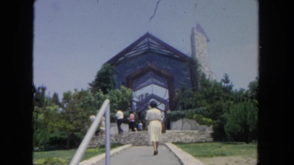 A handheld 8mm view follows the woman from behind as she nears the steps and visitors at the glass-and-stone building.