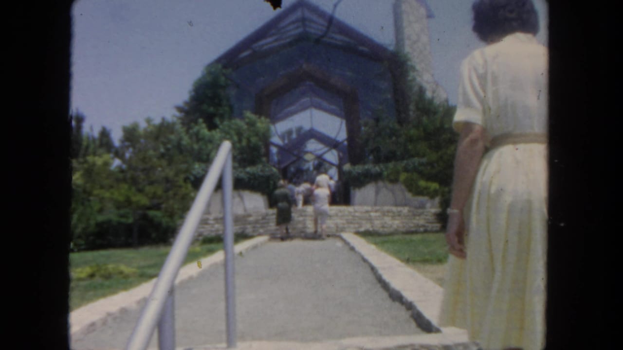 Woman in a white dress walks up a path toward stone steps and a glass-fronted structure.