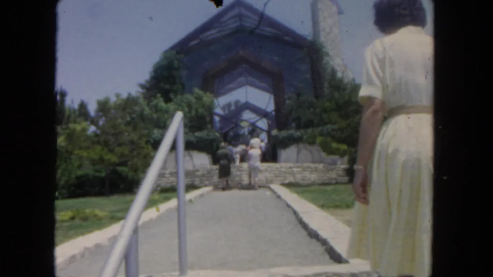 1962 San Diego home-movie frame showing a woman from behind walking up a landscaped path toward a modern glass-fronted structure.