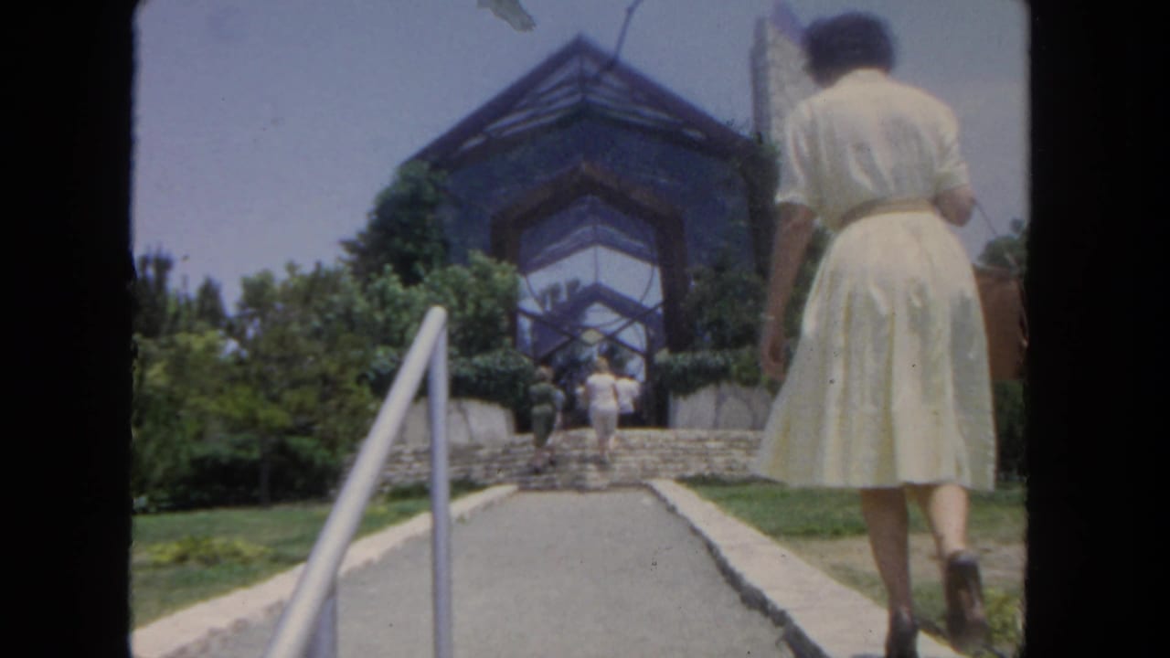 1962 San Diego frame with a woman in a white dress and handbag walking toward stone steps beneath a clear sky.