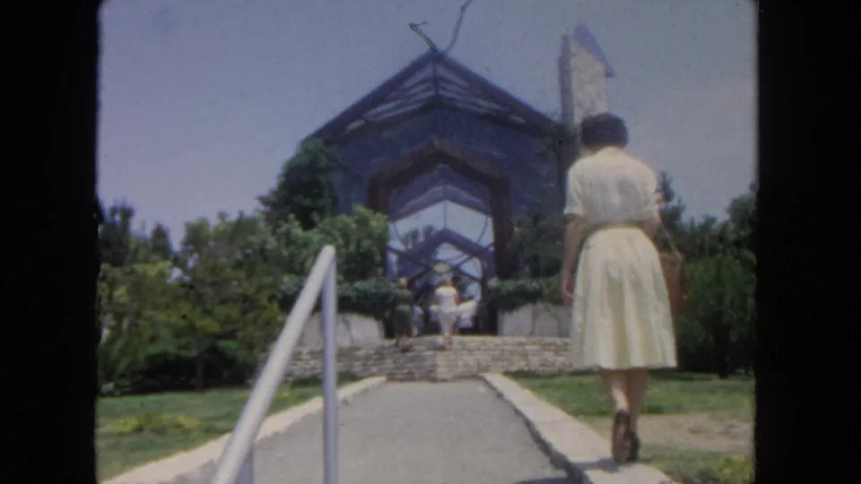 1962 San Diego home-movie frame showing a rear view of a woman walking toward a glass-and-steel structure over stone steps.