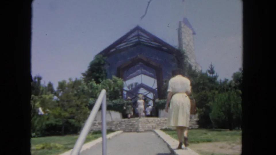 1962 San Diego frame showing the woman near landscaped steps leading to a modern glass-fronted entrance with visitors ahead.